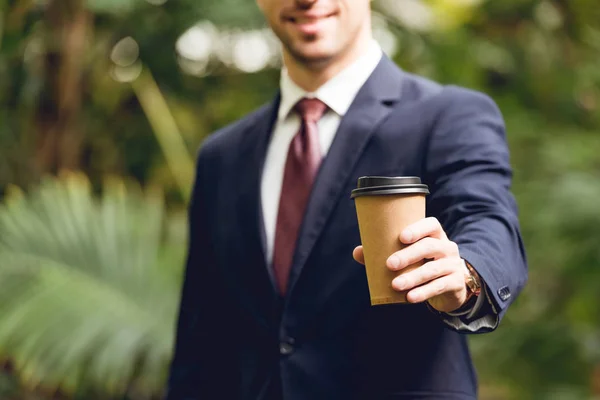 Vista recortada de hombre de negocios sonriente en traje y corbata sosteniendo café para ir en naranjería - foto de stock