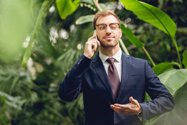 Schöner Geschäftsmann in Anzug, Krawatte und Brille mit geschlossenen Augen, der mit dem Smartphone spricht und im Gewächshaus frische Luft atmet — Stockfoto