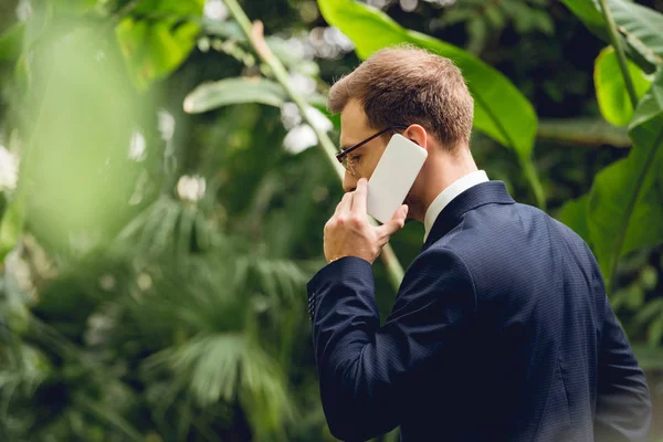 Businessman in suit and glasses talking on smartphone in greenhouse — Stock Photo