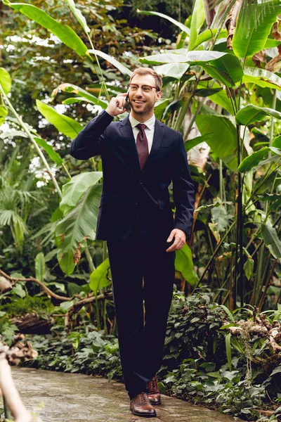 Happy businessman in suit and glasses talking on smartphone while walking in greenhouse — Stock Photo