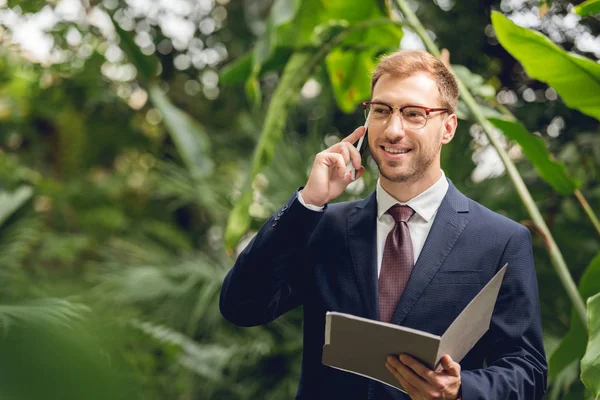 Homme d'affaires heureux en costume et lunettes parlant sur smartphone et tenant dossier en serre — Photo de stock