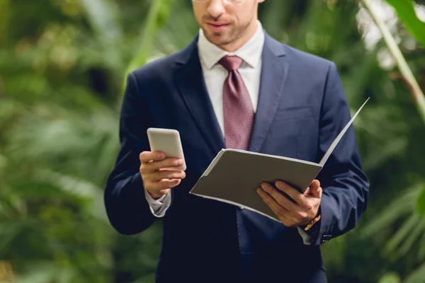 Cropped view of businessman in suit using smartphone and holding folder in greenhouse — Stock Photo