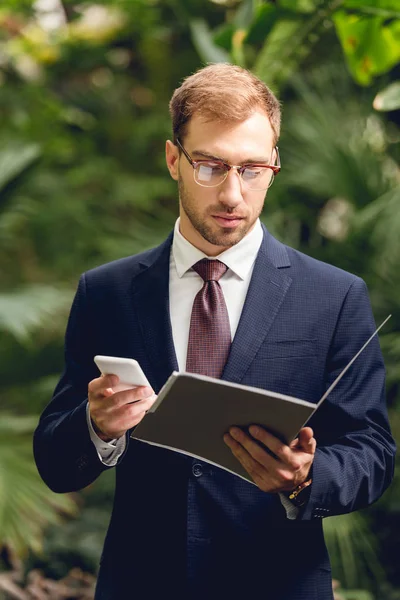 Serious businessman in suit using smartphone and holding folder in greenhouse — Stock Photo