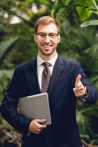 Hombre de negocios feliz en traje y gafas que sostienen la carpeta y señalan con el dedo a la cámara en invernadero - foto de stock