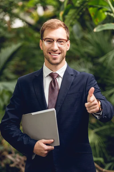 Glücklicher Geschäftsmann in Anzug und Brille, Ordner in der Hand und mit dem Finger auf Kamera im Gewächshaus zeigend — Stockfoto