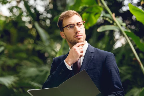 Dreamy businessman in suit and glasses holding folder in greenhouse — Stock Photo