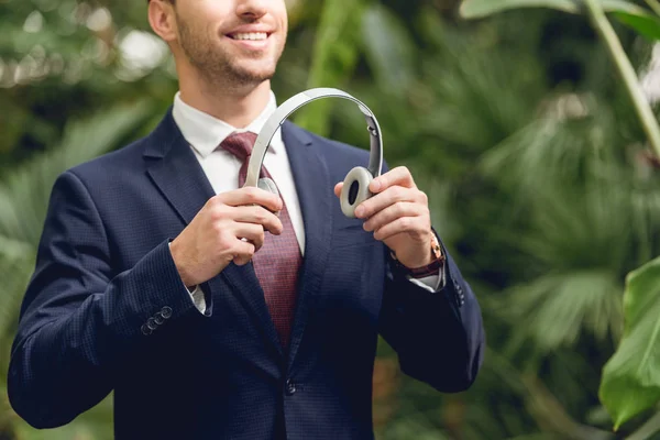 Recortado vista de sonriente hombre de negocios en traje y corbata celebración de auriculares inalámbricos en invernadero - foto de stock