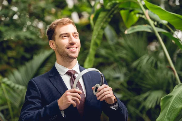 Smiling businessman in suit and tie holding wireless headphones in greenhouse — Stock Photo