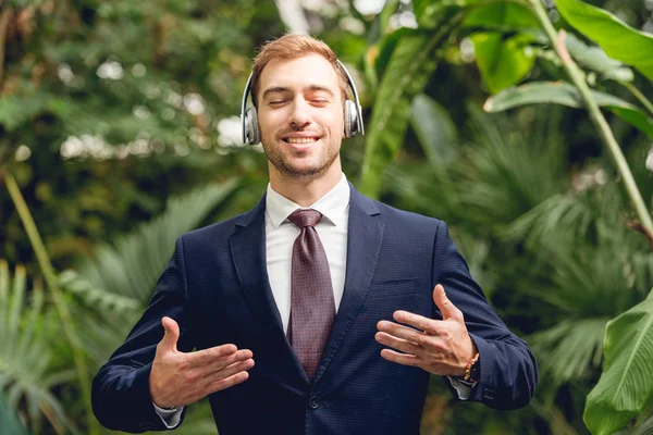 Hombre de negocios feliz en traje y auriculares inalámbricos respirando aire fresco en invernadero - foto de stock