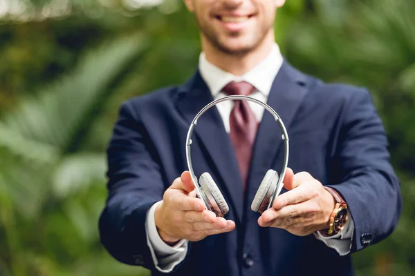 Partial view of smiling businessman in suit and tie holding wireless headphones in greenhouse — Stock Photo