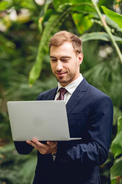 Lächelnder Geschäftsmann in Anzug und Krawatte mit Laptop in Orangerie — Stockfoto