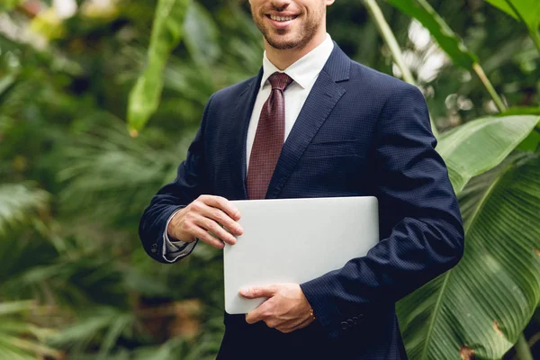 Recortado vista de sonriente hombre de negocios en traje y corbata celebración de ordenador portátil en naranja - foto de stock