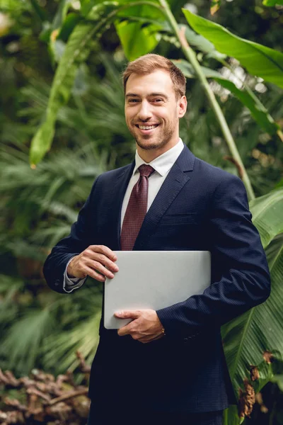 Sonriente hombre de negocios con traje y corbata sosteniendo portátil en naranjería — Stock Photo