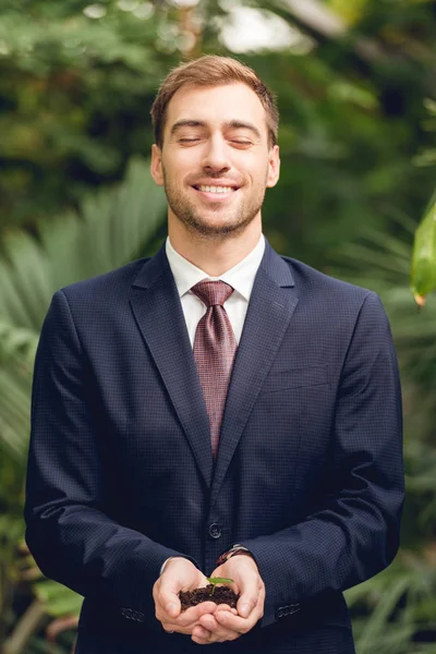 Feliz hombre de negocios sonriente con los ojos cerrados en traje y corbata sosteniendo brote verde y tierra en las manos en naranjería - foto de stock
