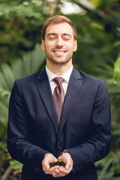 Happy smiling businessman with closed eyes in suit and tie holding green sprout and ground in hands in orangery — Stock Photo