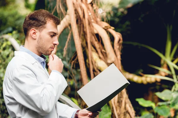 Pensive scientist in white coat reading book in orangery — Stock Photo