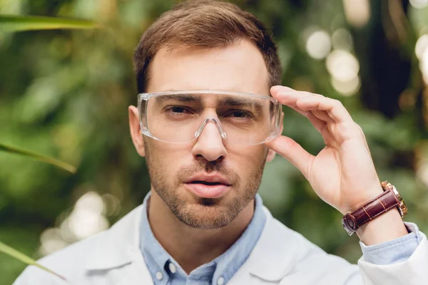 Thoughtful scientist in white coat and goggles in green orangery — Stock Photo