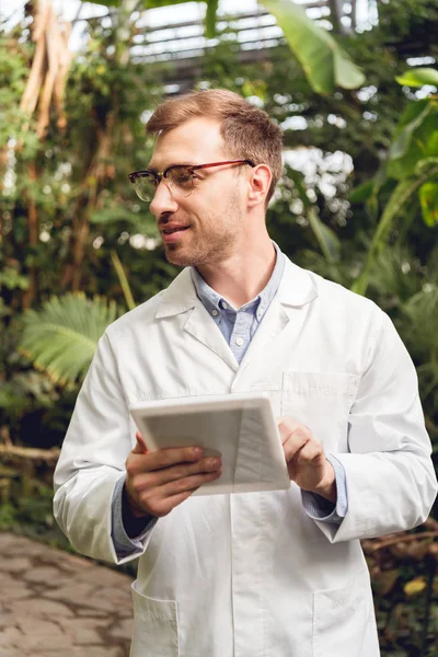 Smiling handsome scientist in white coat and glasses using digital tablet in orangery — Stock Photo