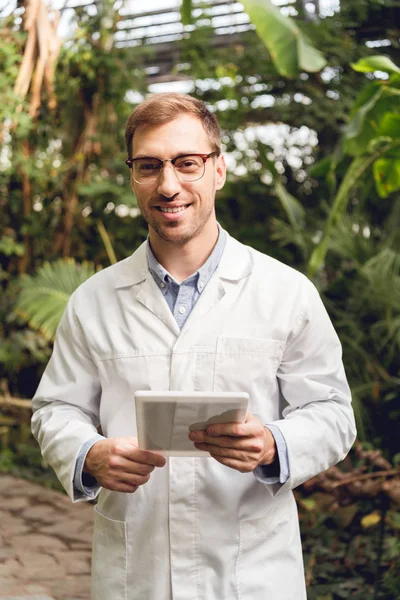 Smiling handsome scientist in white coat and glasses holding digital tablet in orangery — Stock Photo