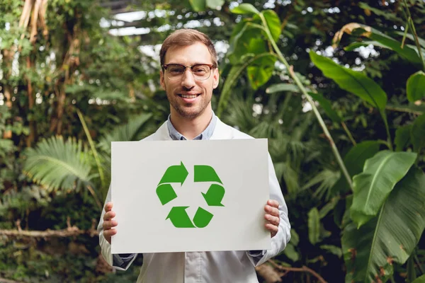 Científico guapo sonriente en bata blanca y gafas con tarjeta con cartel de reciclaje verde en naranja - foto de stock