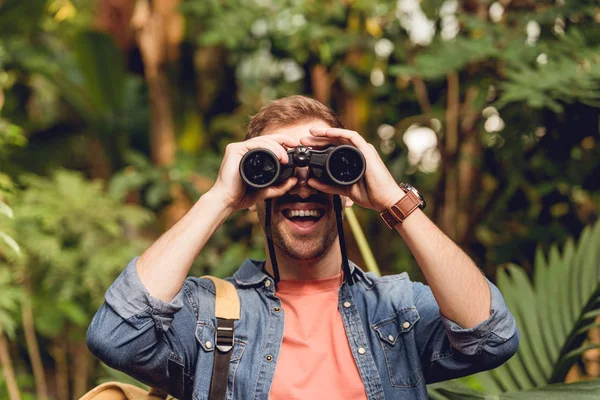 Adulto feliz viajero mirando a través de prismáticos en tropical verde bosque - foto de stock