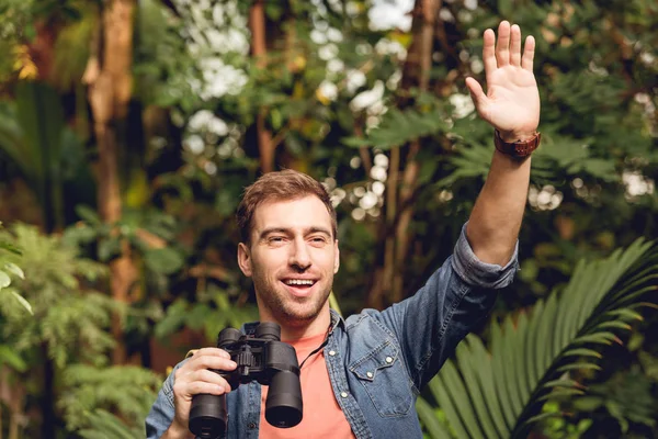 Adult happy traveler with hand up holding binoculars in tropical green forest — Stock Photo