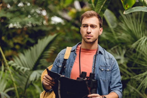 Guapo viajero tenso con prismáticos y mochila sosteniendo mapa en verde bosque tropical - foto de stock