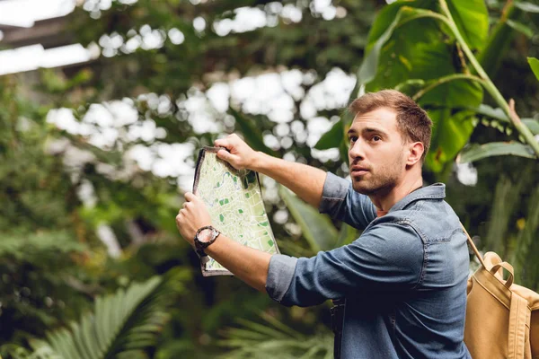 Guapo viajero con mochila sosteniendo mapa en verde bosque tropical - foto de stock