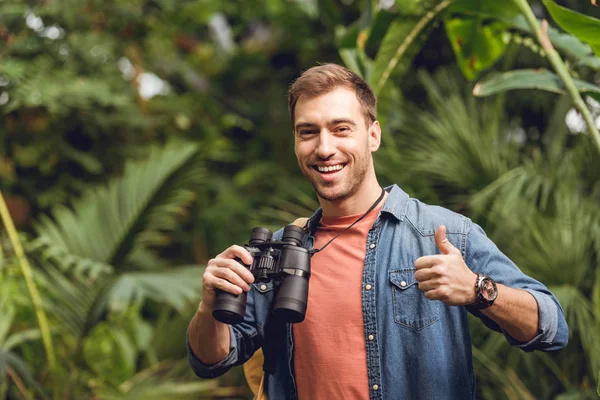 Bonito sorrindo viajante com binóculos e mochila mostrando polegar para cima na floresta tropical verde — Fotografia de Stock