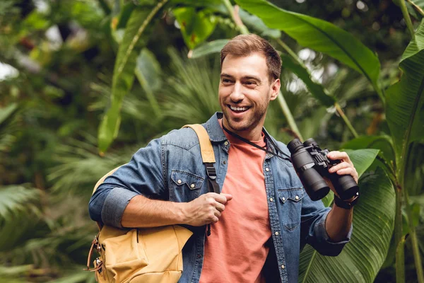 Красивый улыбающийся путешественник с биноклем и рюкзаком в зеленом тропическом лесу — стоковое фото