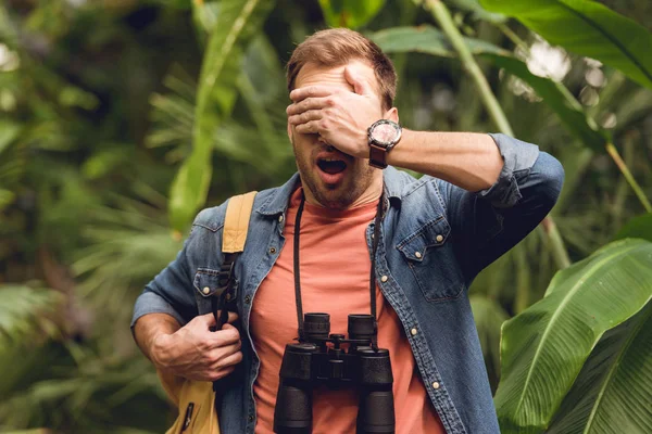 Handsome scared traveler with binoculars and backpack putting hand on eyes in green tropical forest — Stock Photo