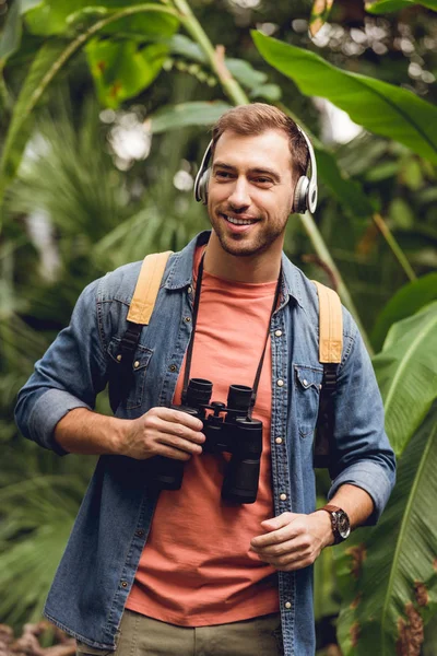 Happy traveler with backpack and binoculars listening music in headphones in green tropical forest — Stock Photo