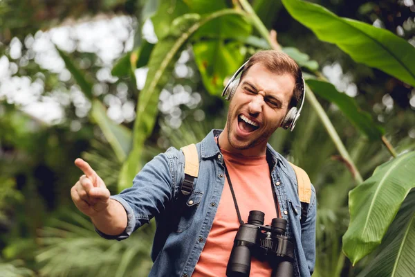 Excited traveler with backpack and binoculars listening music in headphones and singing in tropical forest — Stock Photo