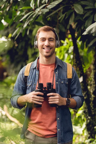 Smiling traveler with backpack and binoculars listening music in headphones in green tropical forest — Stock Photo