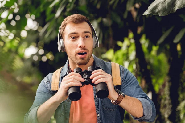 Viaggiatore sorpreso con zaino e binocolo ascoltare musica in cuffia nella foresta tropicale verde — Foto stock