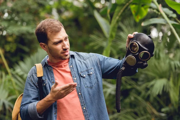 Confused traveler with backpack pointing at gas mask in tropical forest — Stock Photo