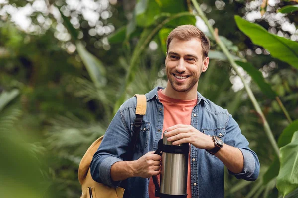 Selective focus of smiling traveler with backpack opening thermos in tropical forest — Stock Photo