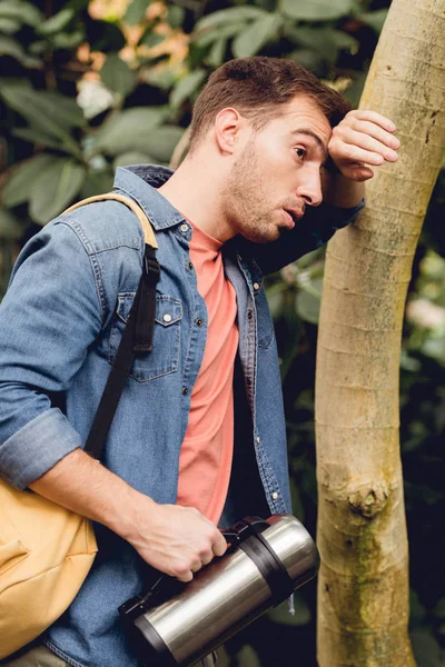 Erschöpfter Reisender mit Rucksack und Thermoskanne steht neben Baumstamm im Tropenwald — Stockfoto