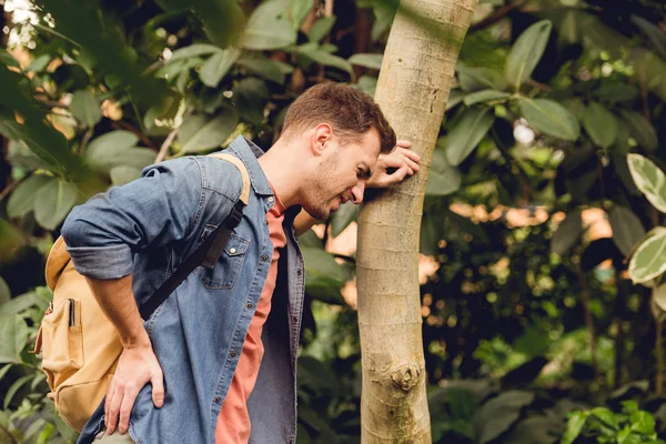 Traveler with backpack and backache standing near tree trunk in tropical forest — Stock Photo