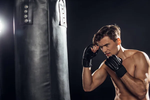 Tired boxer in gloves training with punching bag on black — Stock Photo