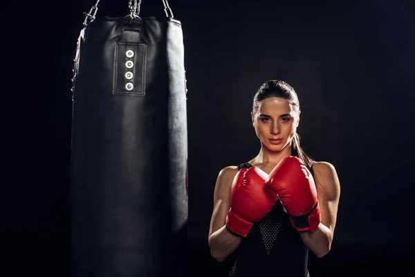 Vista frontale del pugile in guanti da boxe rossi in piedi vicino al sacco da boxe sul nero — Foto stock