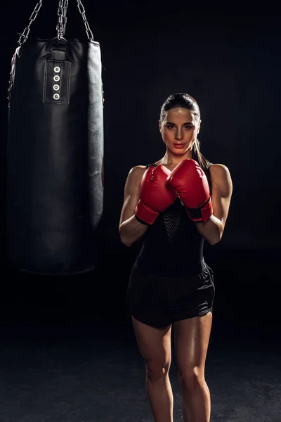 Vista frontal do boxeador em luvas de boxe vermelho em pé perto de saco de perfuração em preto — Fotografia de Stock