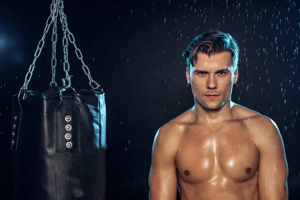 Frontansicht eines Boxers, der in der Nähe eines Boxsacks unter Wassertropfen auf schwarz steht — Stockfoto
