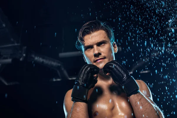 Pensive boxer in leather gloves standing under water drops and looking away — Stock Photo