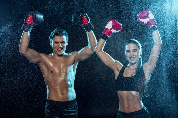 Two boxers in boxing gloves smiling and showing yes gestures under water drops on black — Stock Photo