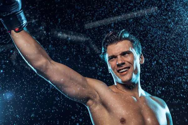 Smiling boxer standing with hand up under water drops on black — Stock Photo