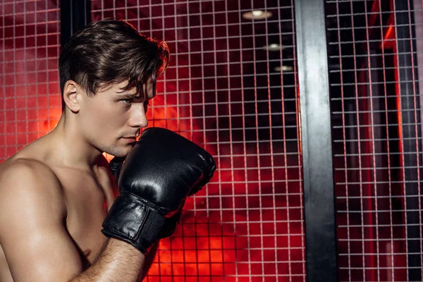 Handsome boxer in boxing gloves standing near wire netting and looking away — Stock Photo