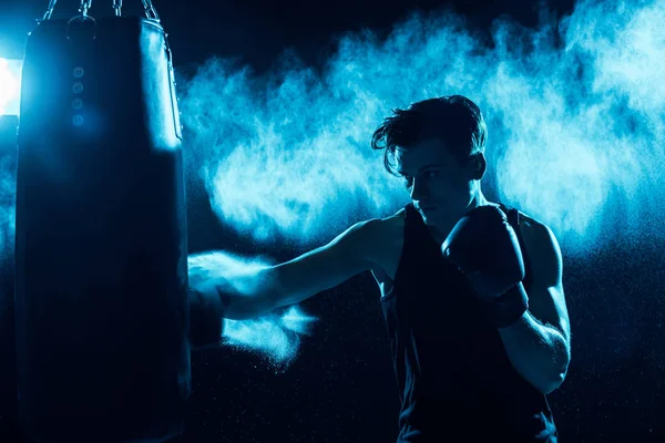 Boxeador concentrado en guantes de boxeo entrenamiento con saco de boxeo en oscuridad - foto de stock