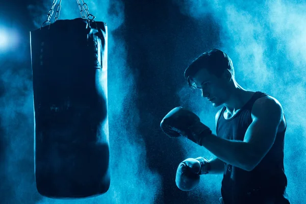 Concentrated boxer in boxing gloves training with punching bag in dark — Stock Photo