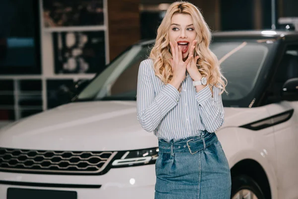 Aufgeregtes blondes Mädchen steht neben weißem Auto in Autosalon — Stockfoto